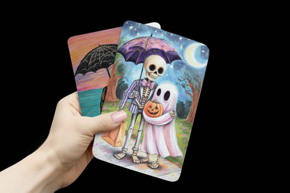 Hand holding tarot cards with Halloween-themed illustrations of a skeleton and ghost couple, card stock, Halloween, seasonal, collection, pink, skeleton family